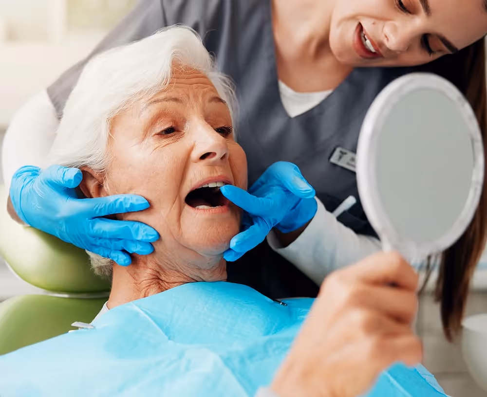 Dentist wearing blue gloves examining an elderly woman's mouth while she looks into a hand mirror.