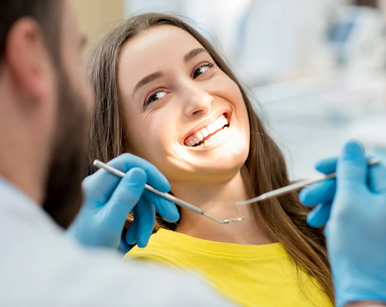 Smiling woman in a yellow shirt during a dental checkup with a dentist holding dental tools.