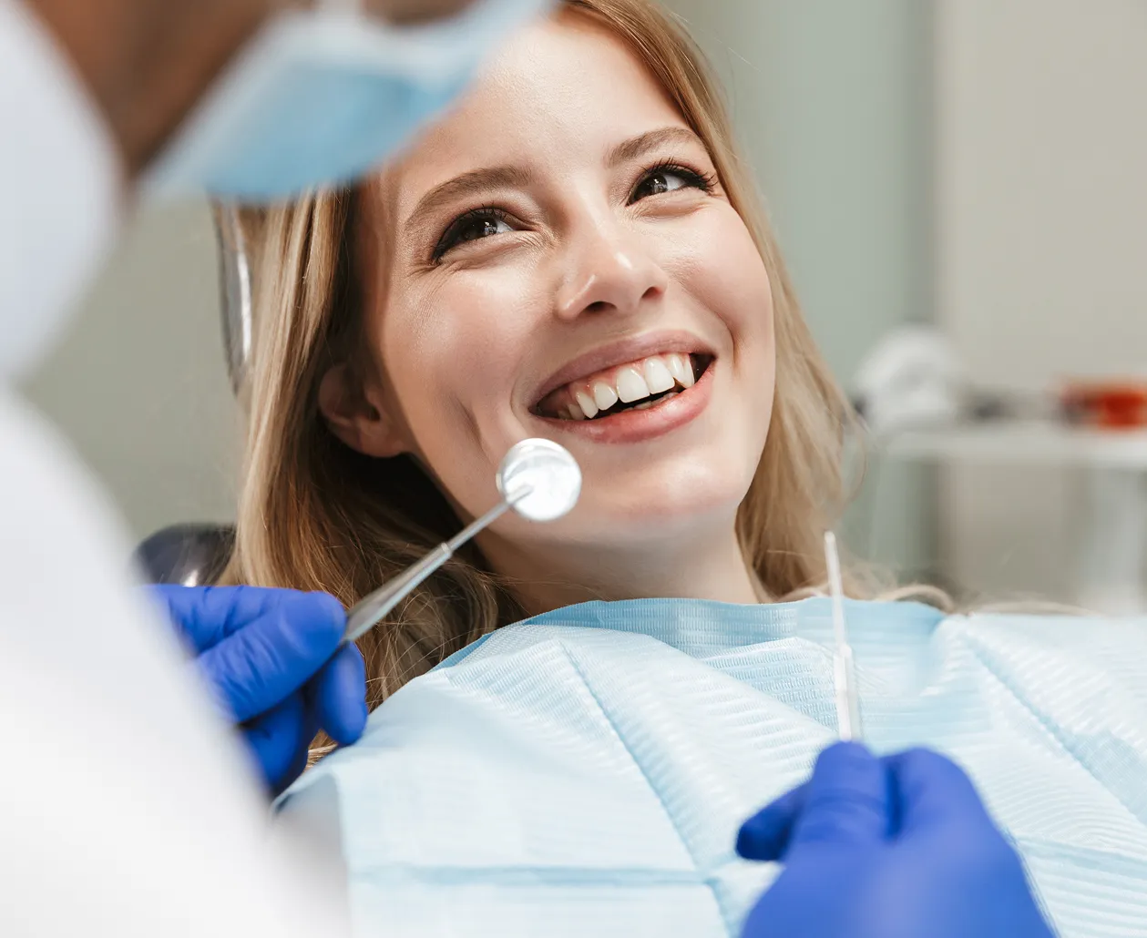 Smiling woman receiving dental checkup from dentist wearing blue gloves and holding dental tools.