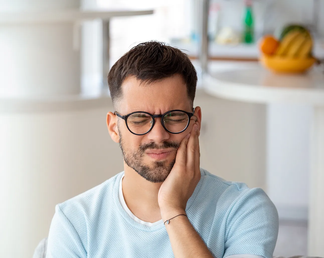 Young man wearing glasses with a beard holding his cheek in pain, indicating toothache or mouth discomfort.
