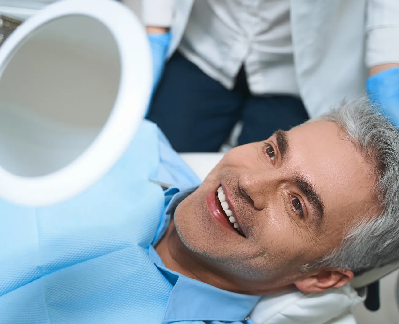 Smiling middle-aged man wearing a blue dental bib looking at a handheld mirror during a dental checkup.