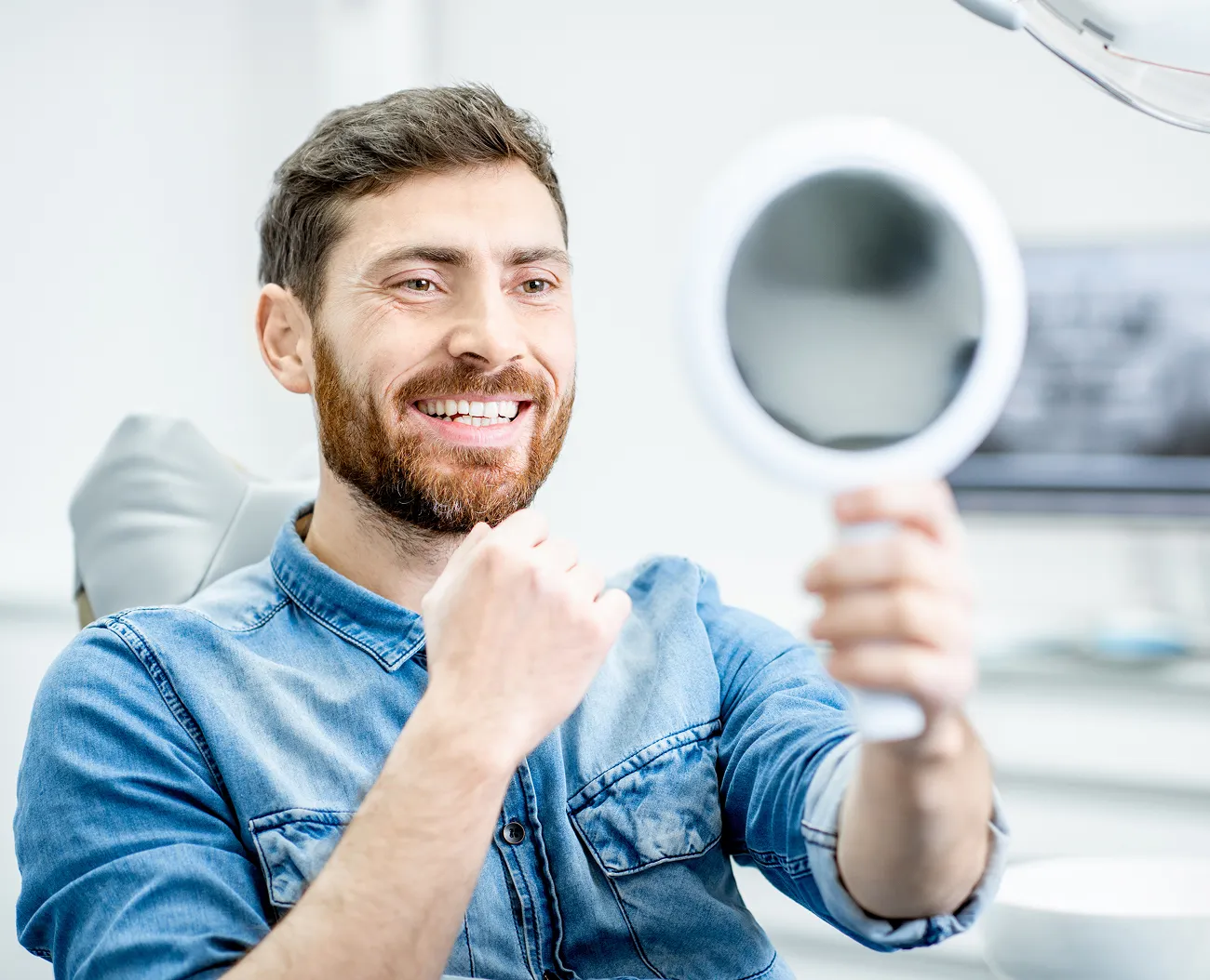 Bearded man wearing a denim shirt smiling and looking at his teeth in a handheld mirror in a dental office.