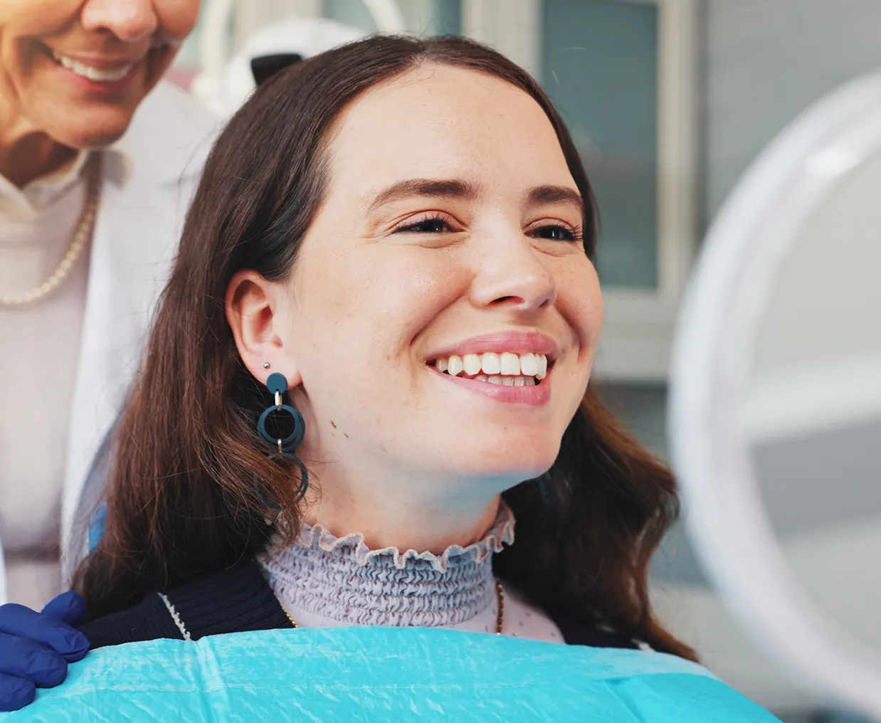 Smiling woman with long brown hair wearing blue earrings looks at her reflection in a mirror while a dental professional in a white coat and gloves stands behind her.