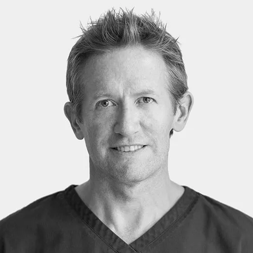 Black and white portrait of a man with short spiky hair wearing a dark V-neck shirt, smiling slightly, against a plain background.