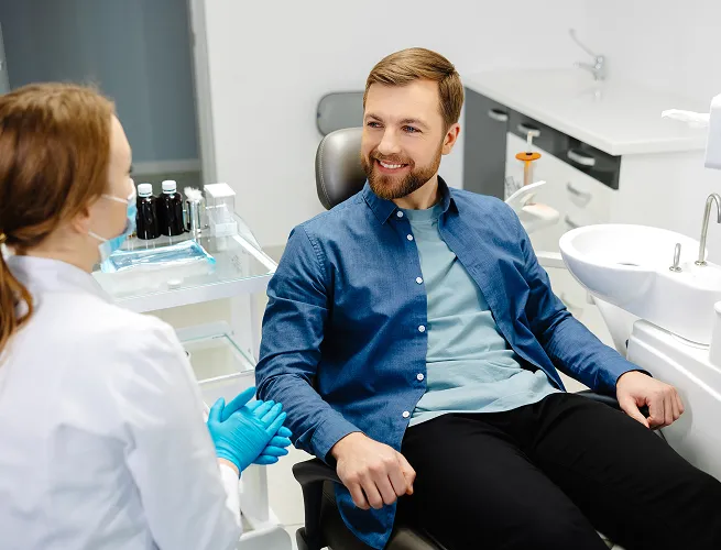 Male patient smiling and sitting in a dental chair while talking to a female dentist wearing gloves and a face mask.