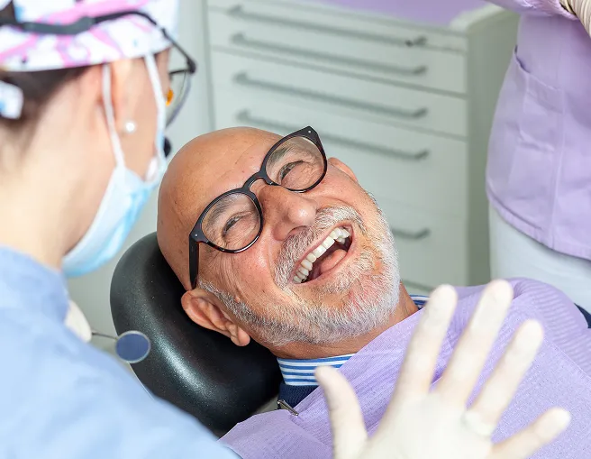 Smiling older man wearing glasses reclining in a dental chair while interacting with a dentist.