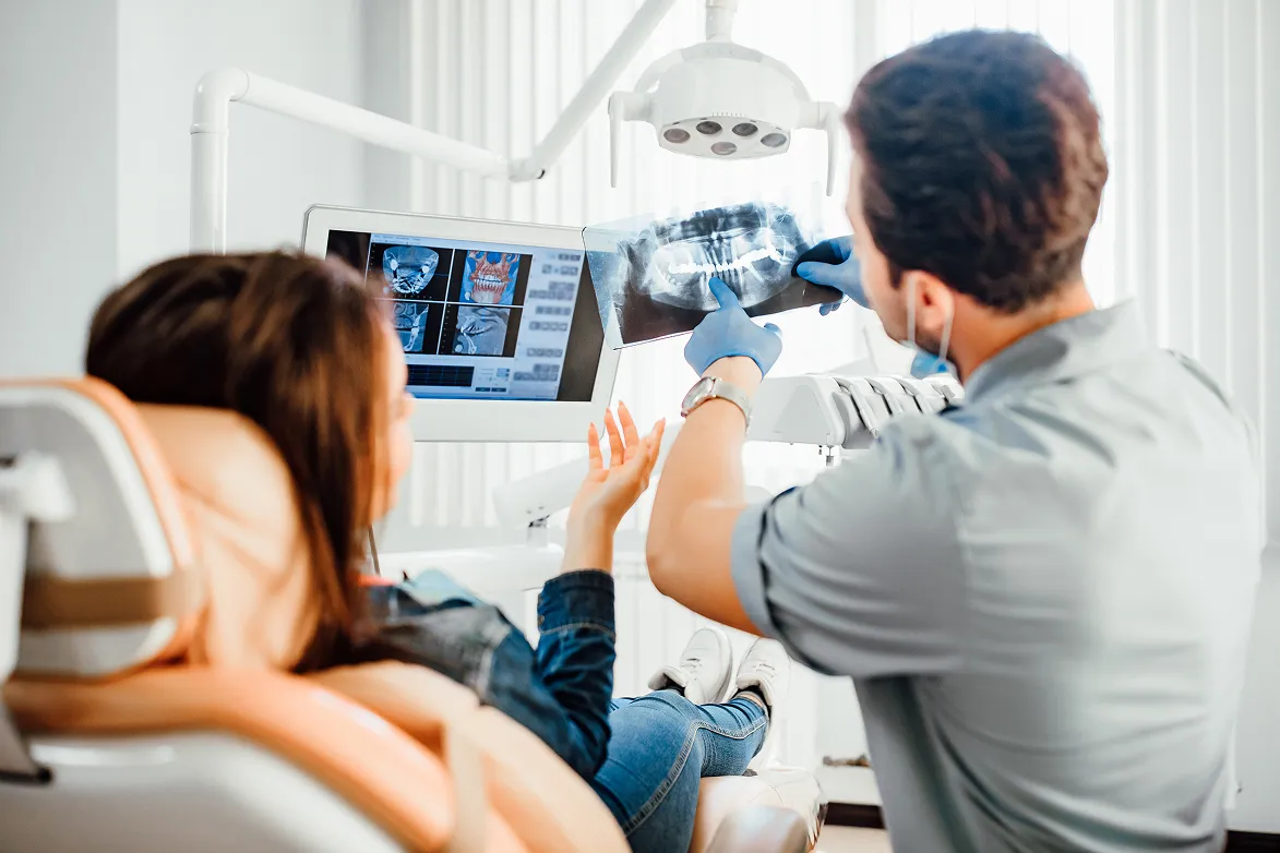 Dentist wearing gloves explaining dental X-ray to female patient reclining in dental chair.