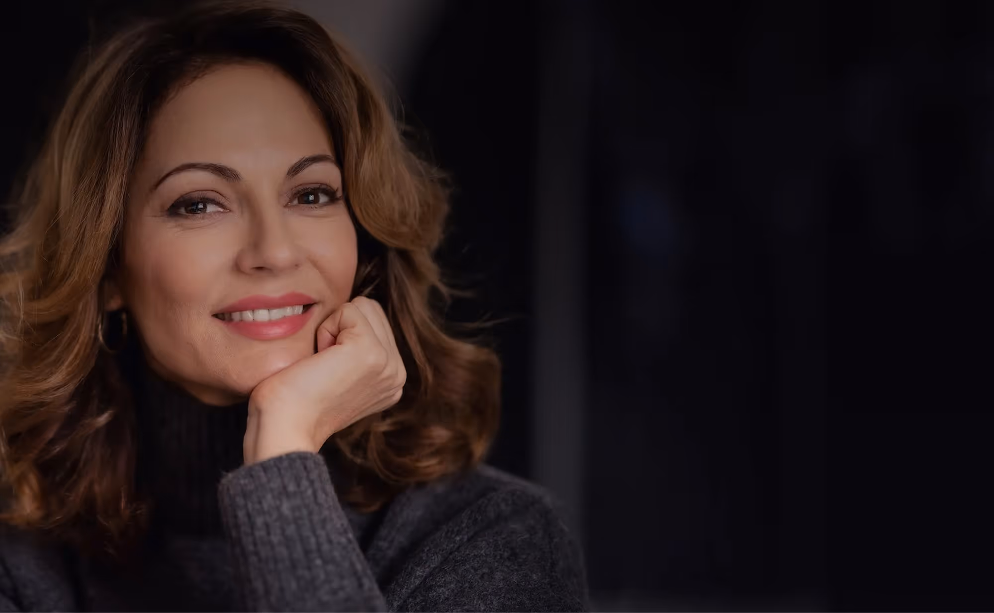 Smiling woman with wavy hair resting her chin on her hand wearing a dark sweater against a dark background.
