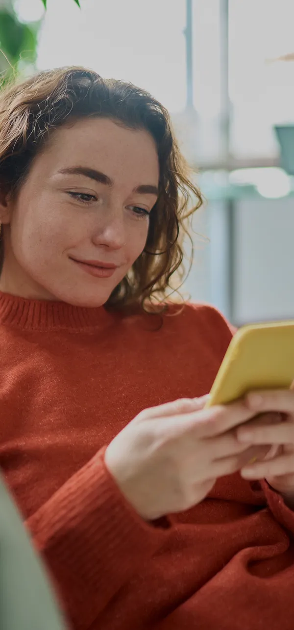 Young woman in a red sweater looking at and holding a yellow smartphone smiling softly.
