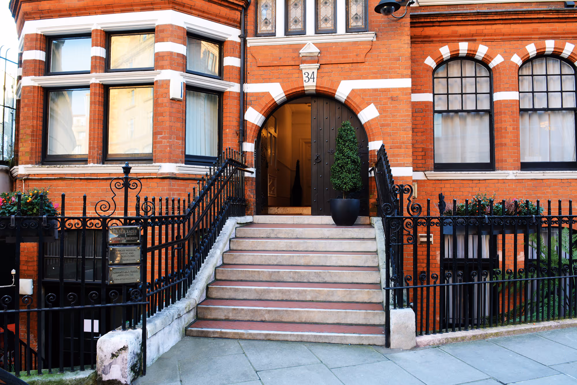 Entrance of a red brick building with black iron railings and a staircase leading to an arched wooden door marked number 34.