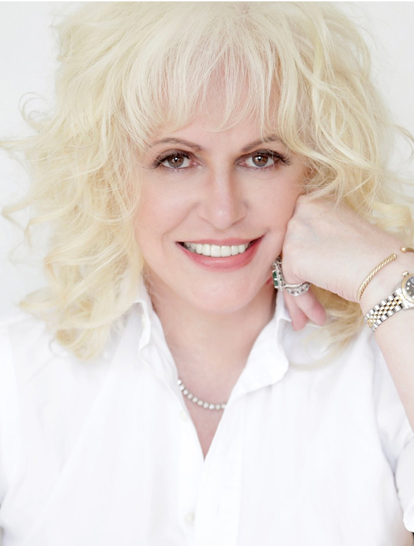 Smiling woman with curly blonde hair resting her head on her fist, wearing a white shirt and jewelry.