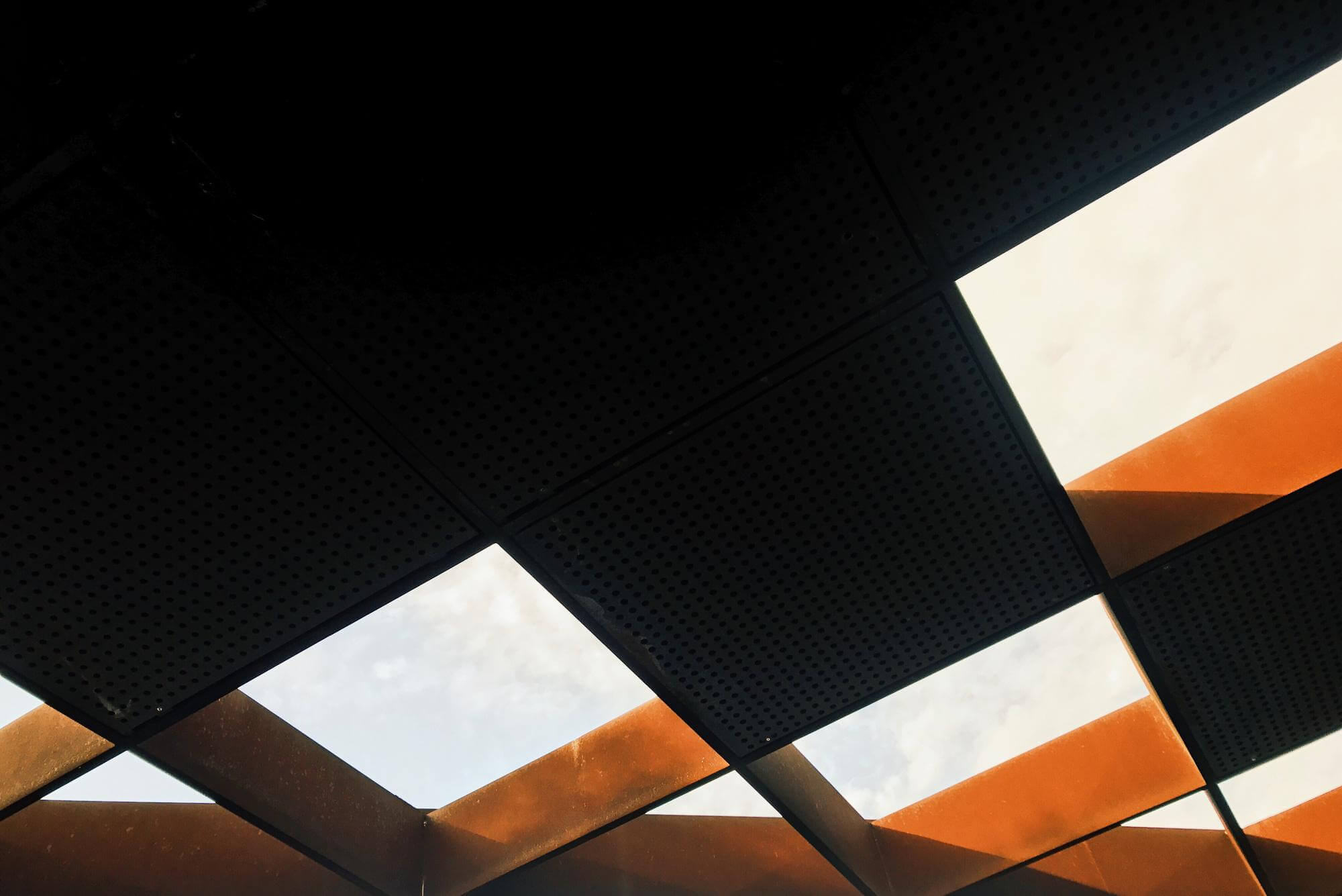 View of a geometric ceiling grid with black perforated panels and orange beams against a cloudy sky.