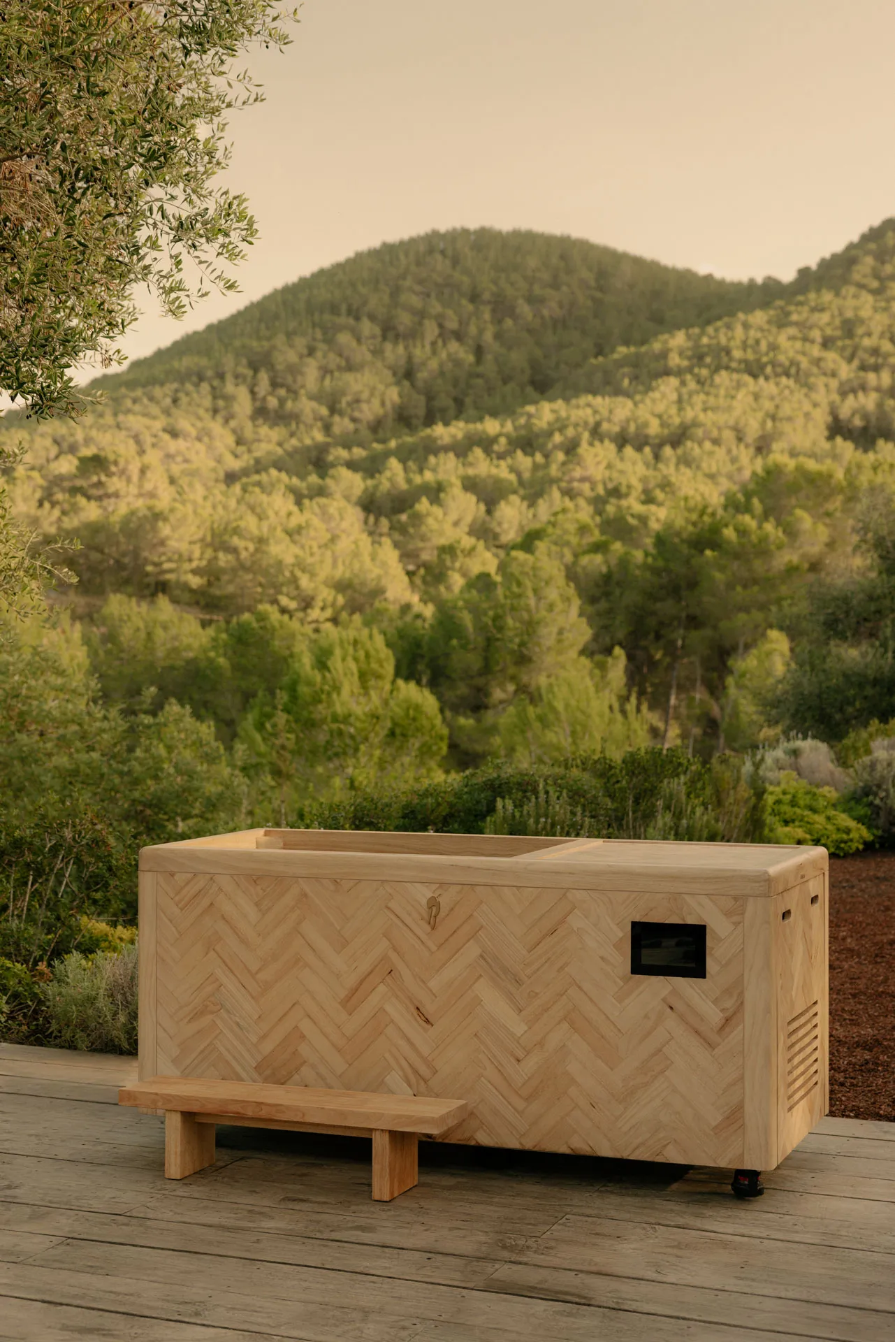 Wooden hot tub with herringbone pattern on a wooden deck overlooking green forested hills during sunset.