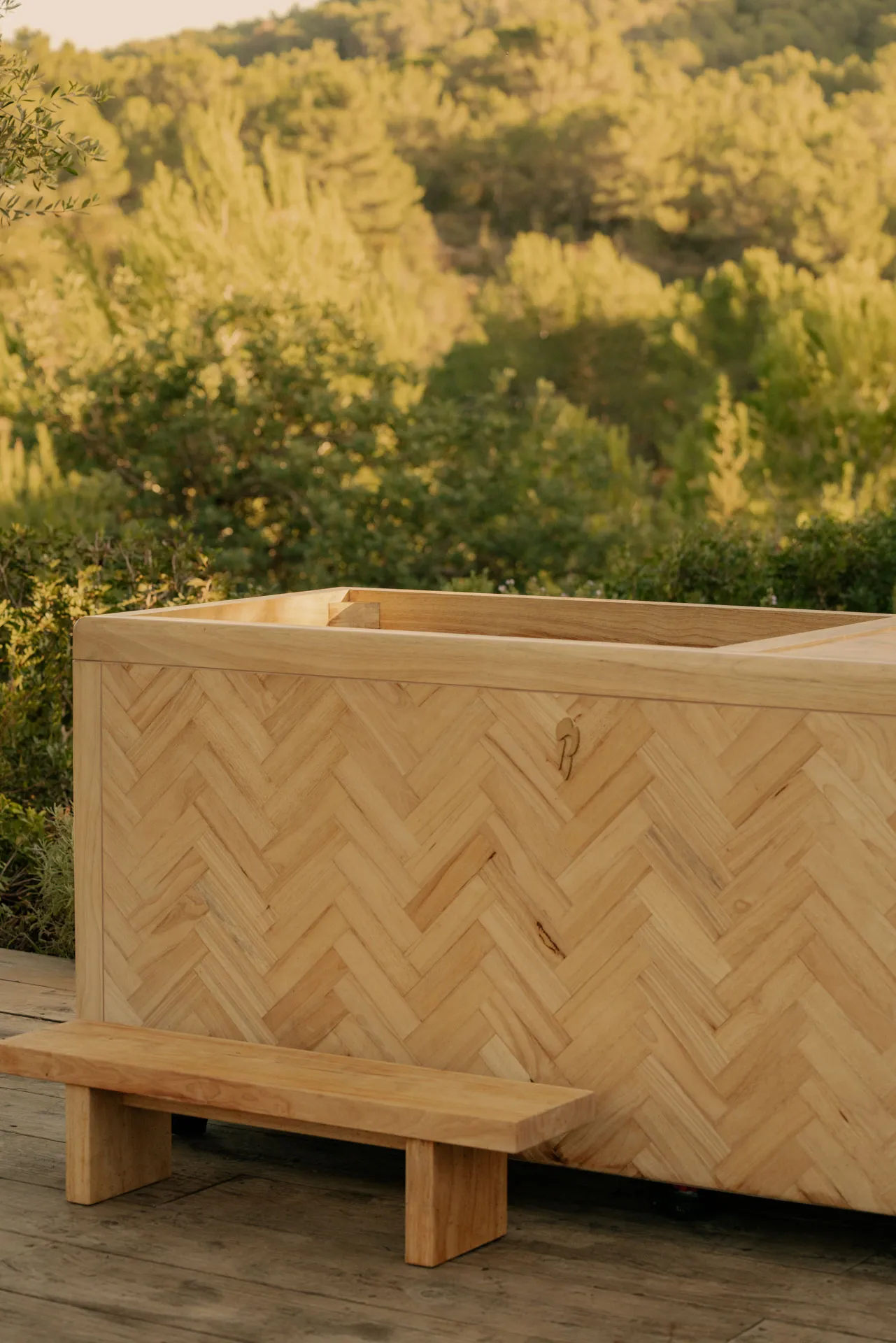 Wooden outdoor hot tub with a herringbone pattern and a small wooden bench on a wooden deck, surrounded by green trees.