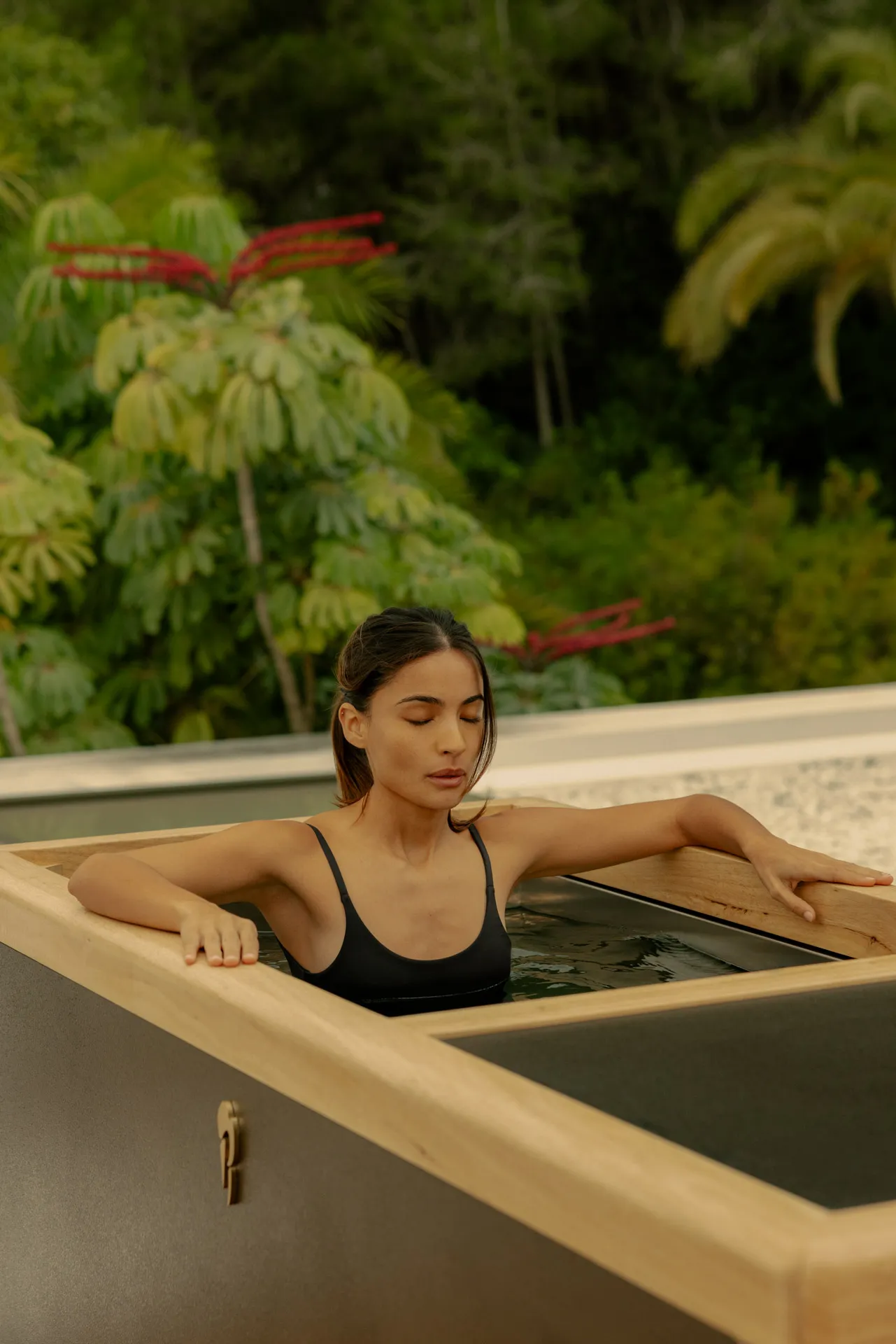 Woman in a black swimsuit relaxing with eyes closed in a wooden hot tub outdoors surrounded by greenery.