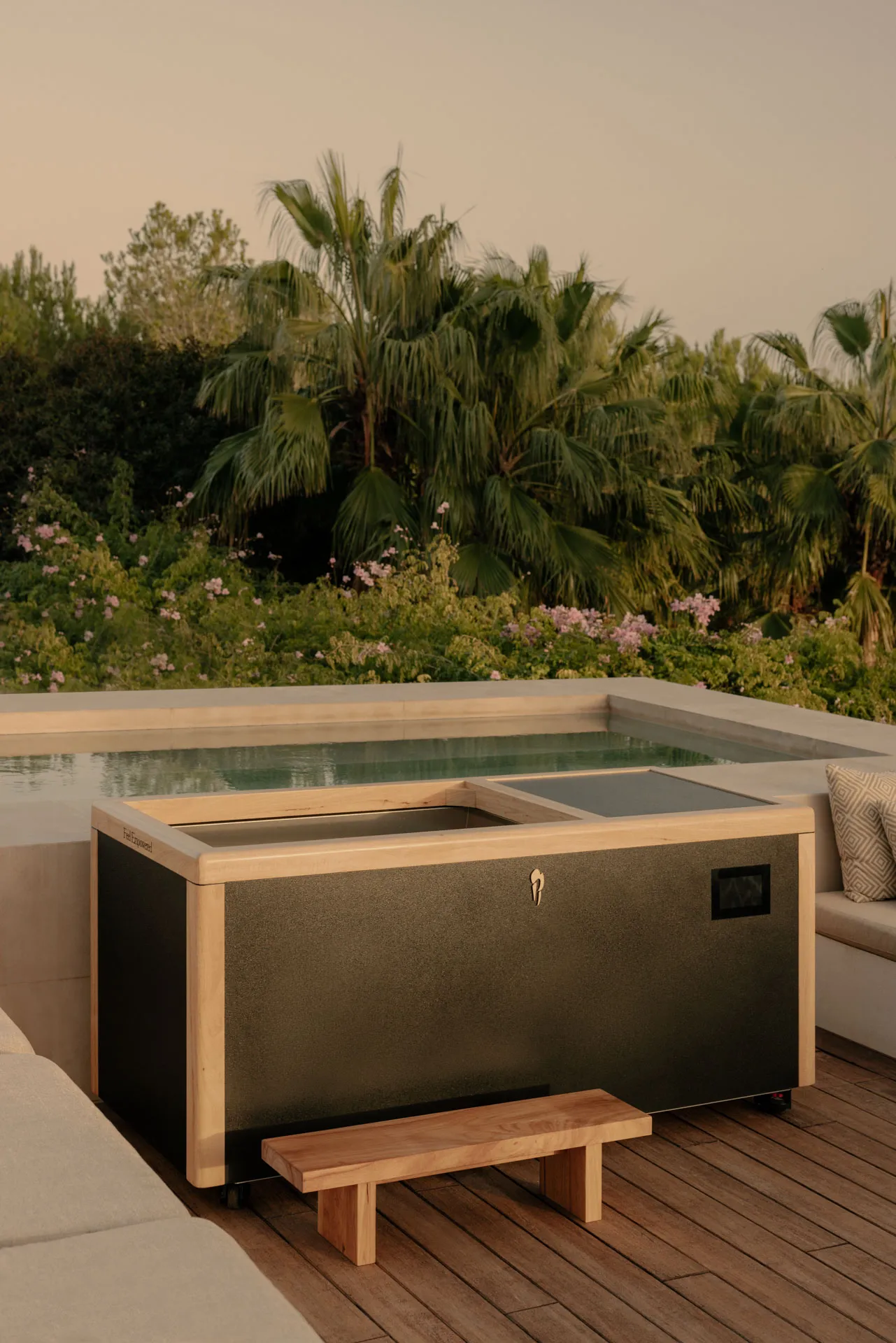 Modern black and wooden hot tub with a small wooden step on a wooden deck surrounded by cushioned seating and tropical plants.