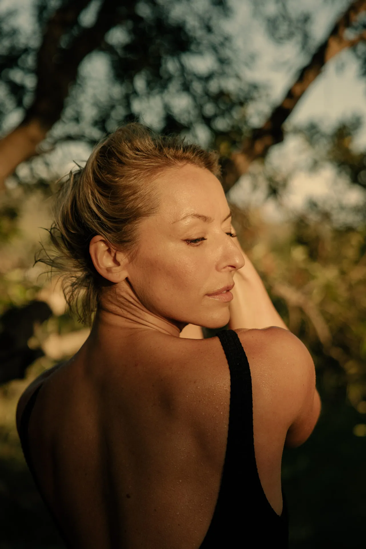Woman with blonde hair in a black sleeveless top, looking over her shoulder outdoors with blurred trees in the background.
