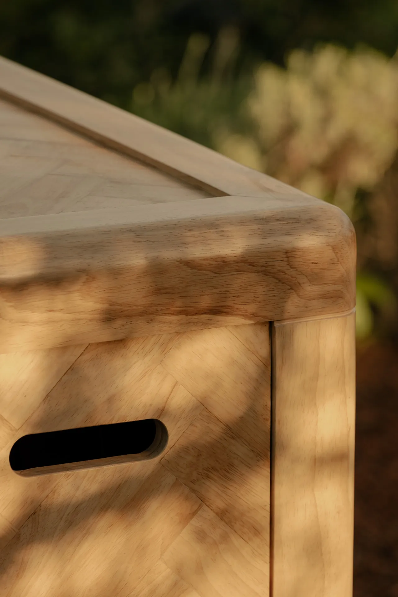 Close-up of a light wooden piece of furniture with smooth rounded edges and a horizontal slot handle, lit by soft natural light.