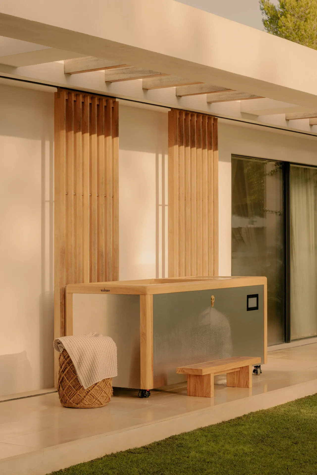 Outdoor wooden hot tub with metal sides on patio next to a woven basket with a folded beige towel and a small wooden step, under a pergola.