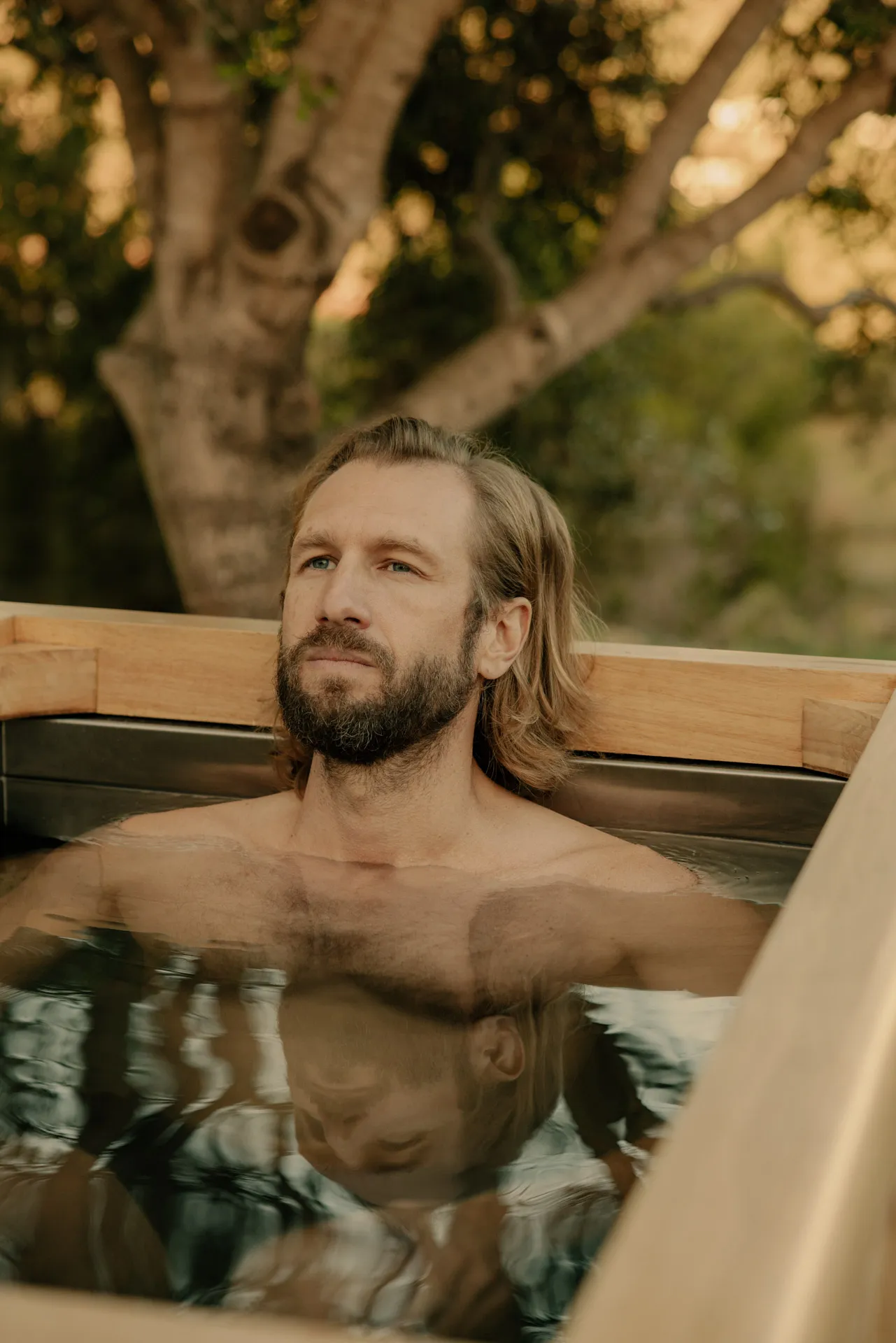 Bearded man with long hair relaxing in an outdoor wooden hot tub with a reflective water surface.