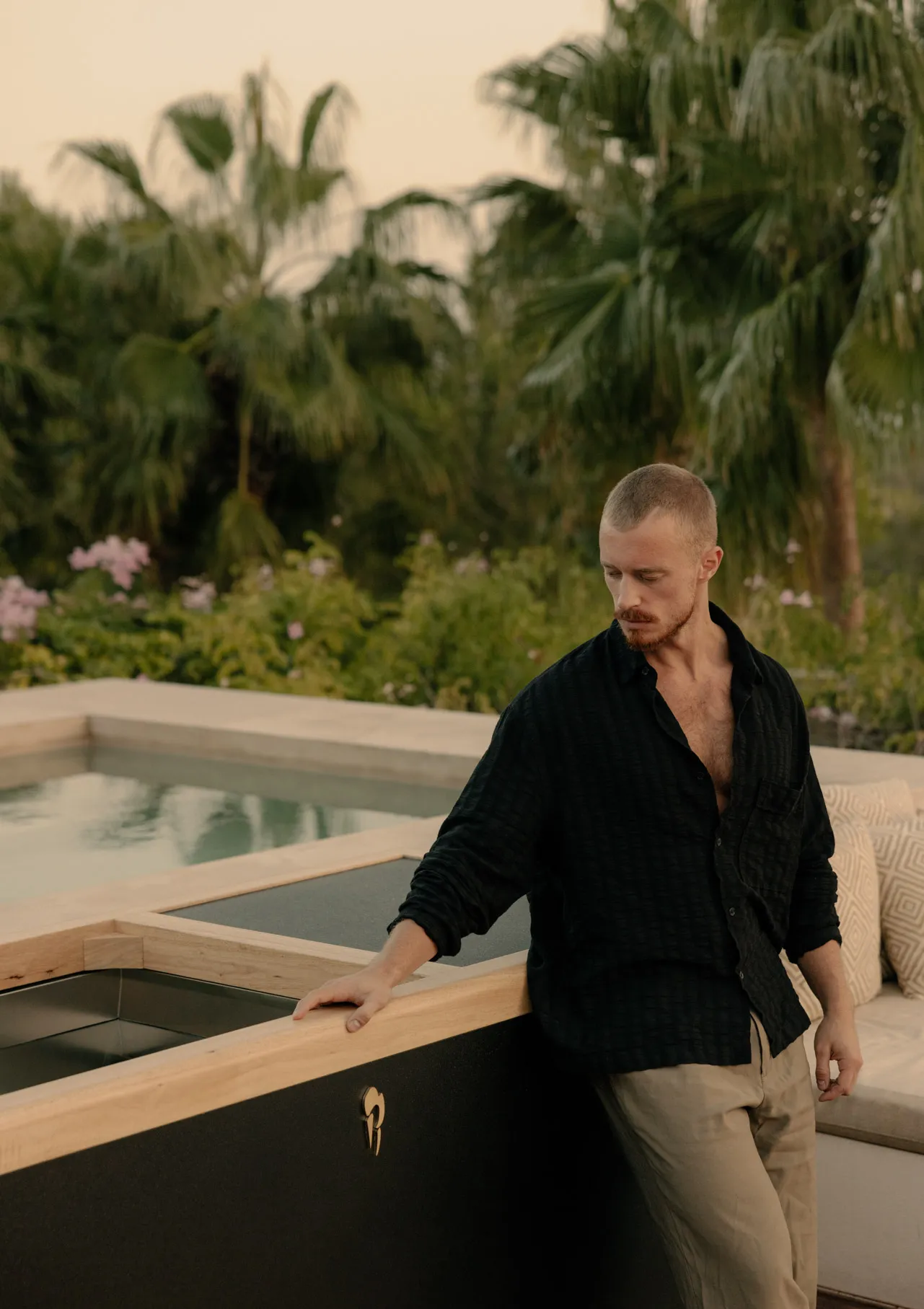 Man in a black shirt and beige pants leaning on a wooden ledge near an outdoor pool with palm trees in the background.