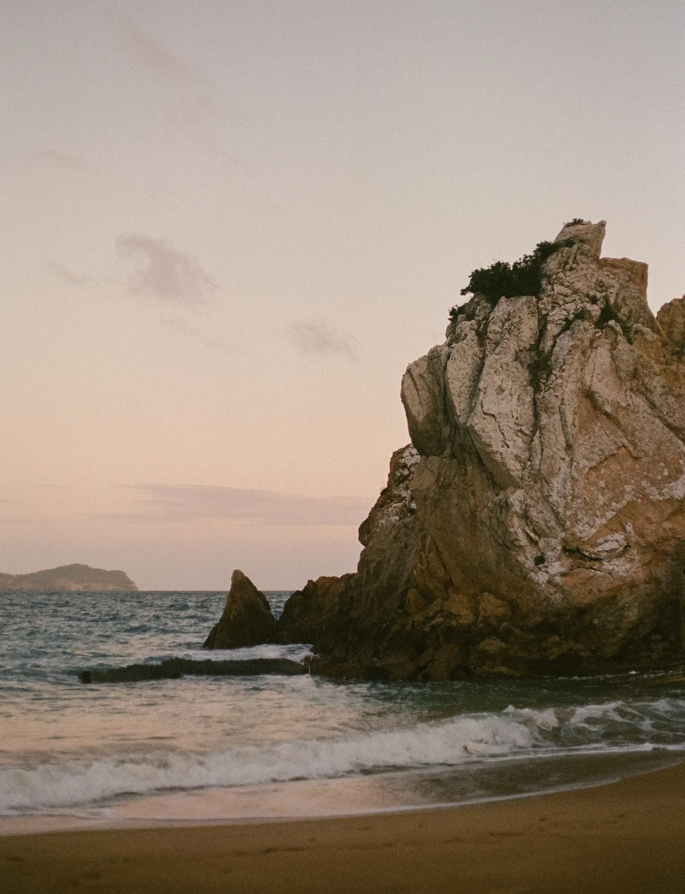 Large rocky cliff on a beach with waves gently crashing on the shore during sunset.