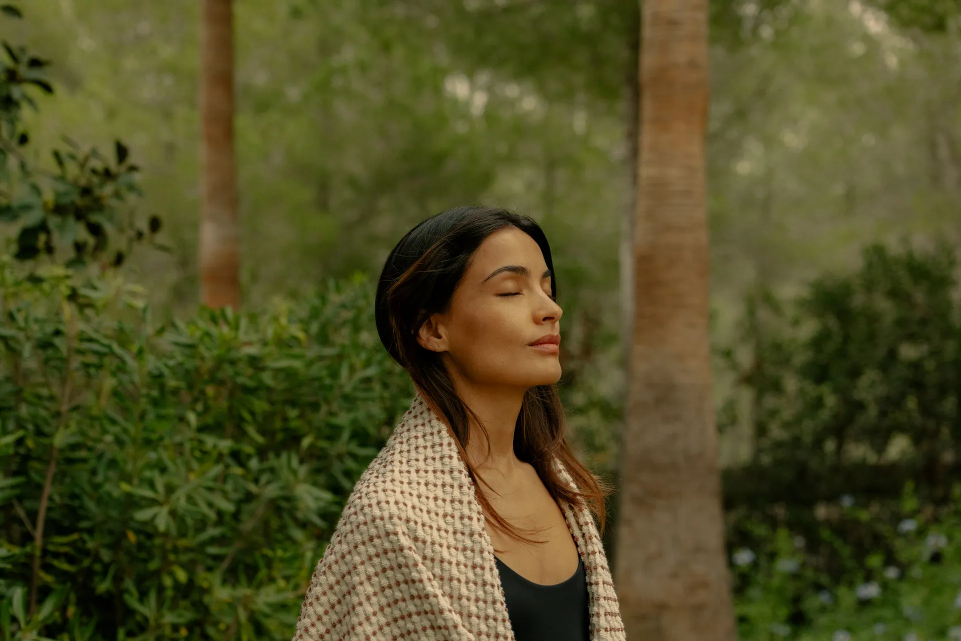 Woman with closed eyes wrapped in a blanket standing outdoors with green foliage and trees in the background.