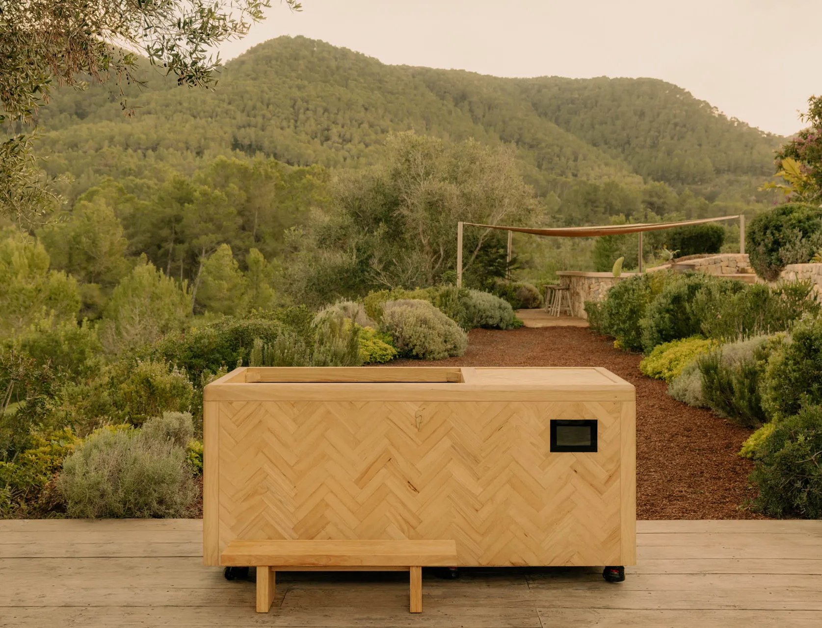 Wooden rectangular planter box on wheels with a small bench in front, surrounded by lush greenery and hills in the background.