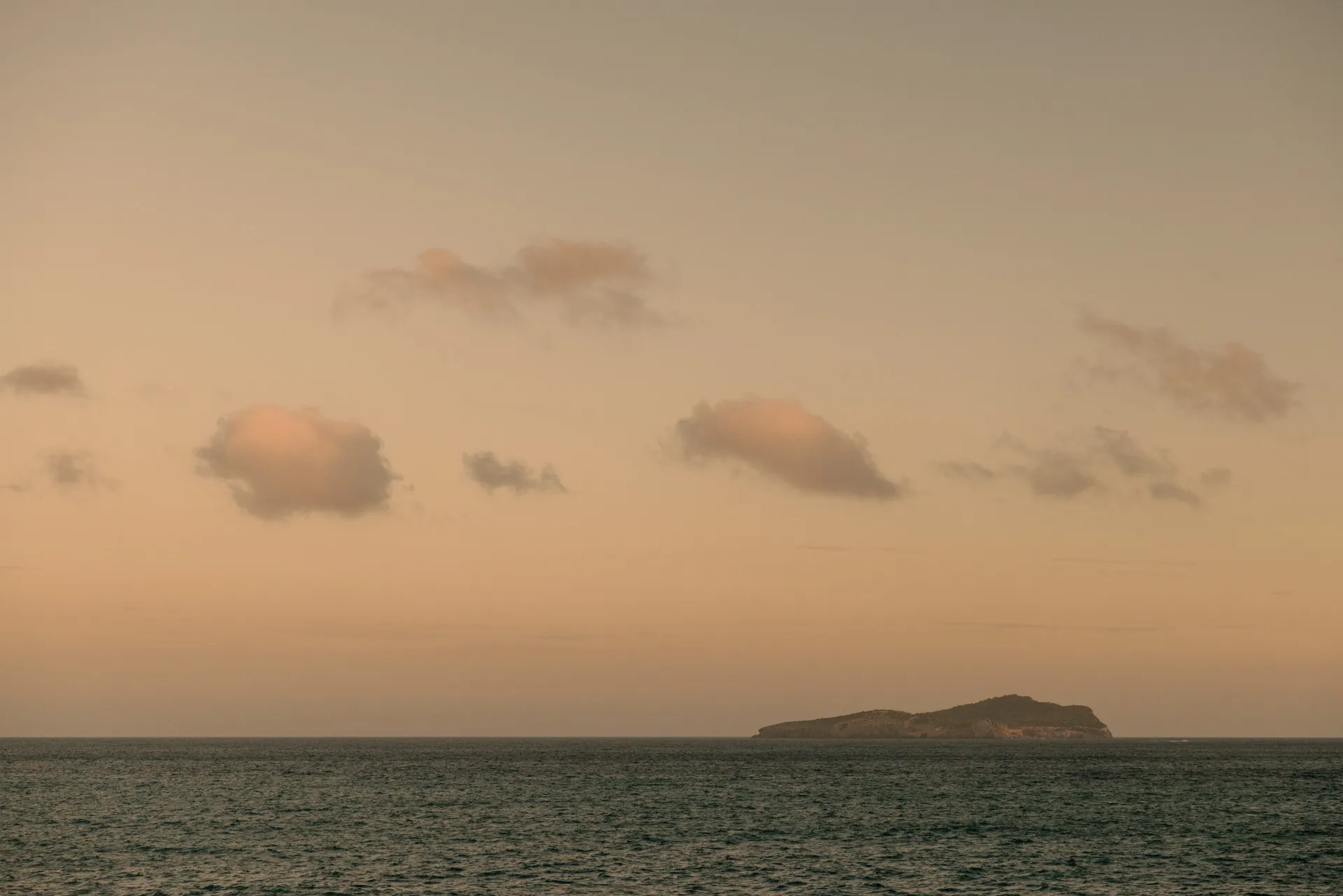 Island silhouette on calm sea under soft orange sky with scattered clouds at sunset.