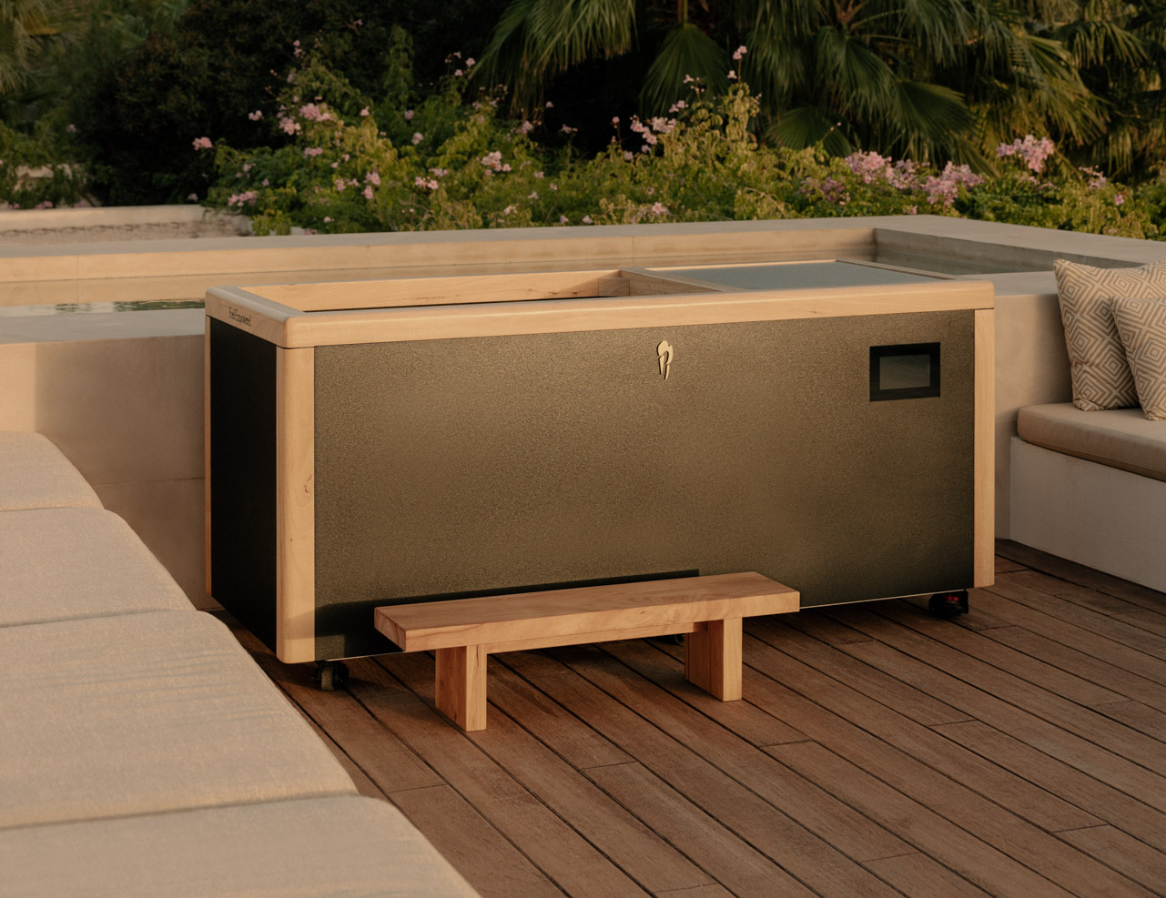 Modern outdoor wooden hot tub on a wooden deck surrounded by cushioned seating and greenery at sunset.