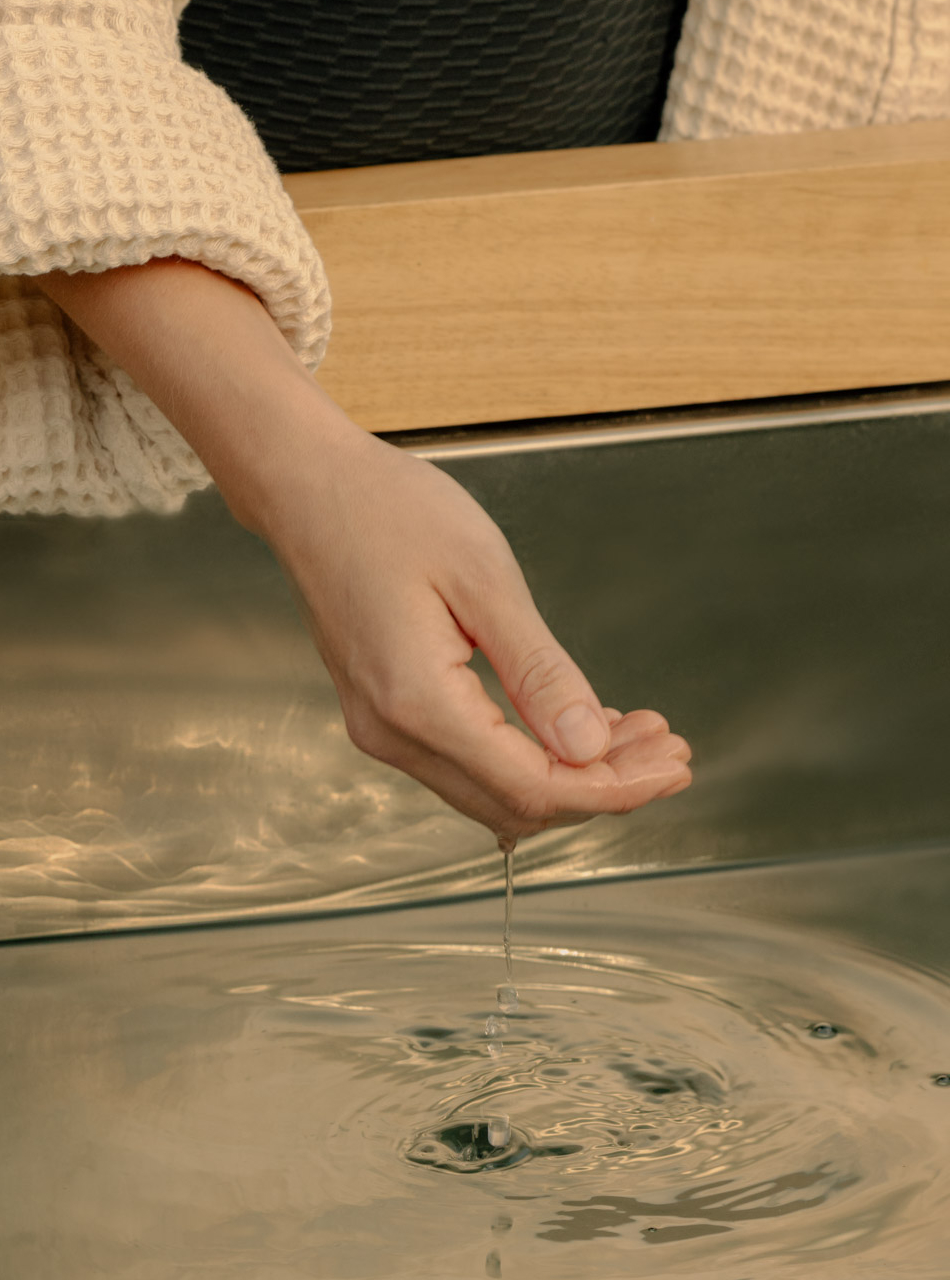 Hand dripping water into a reflective surface creating ripples, with a cream textured sleeve visible.