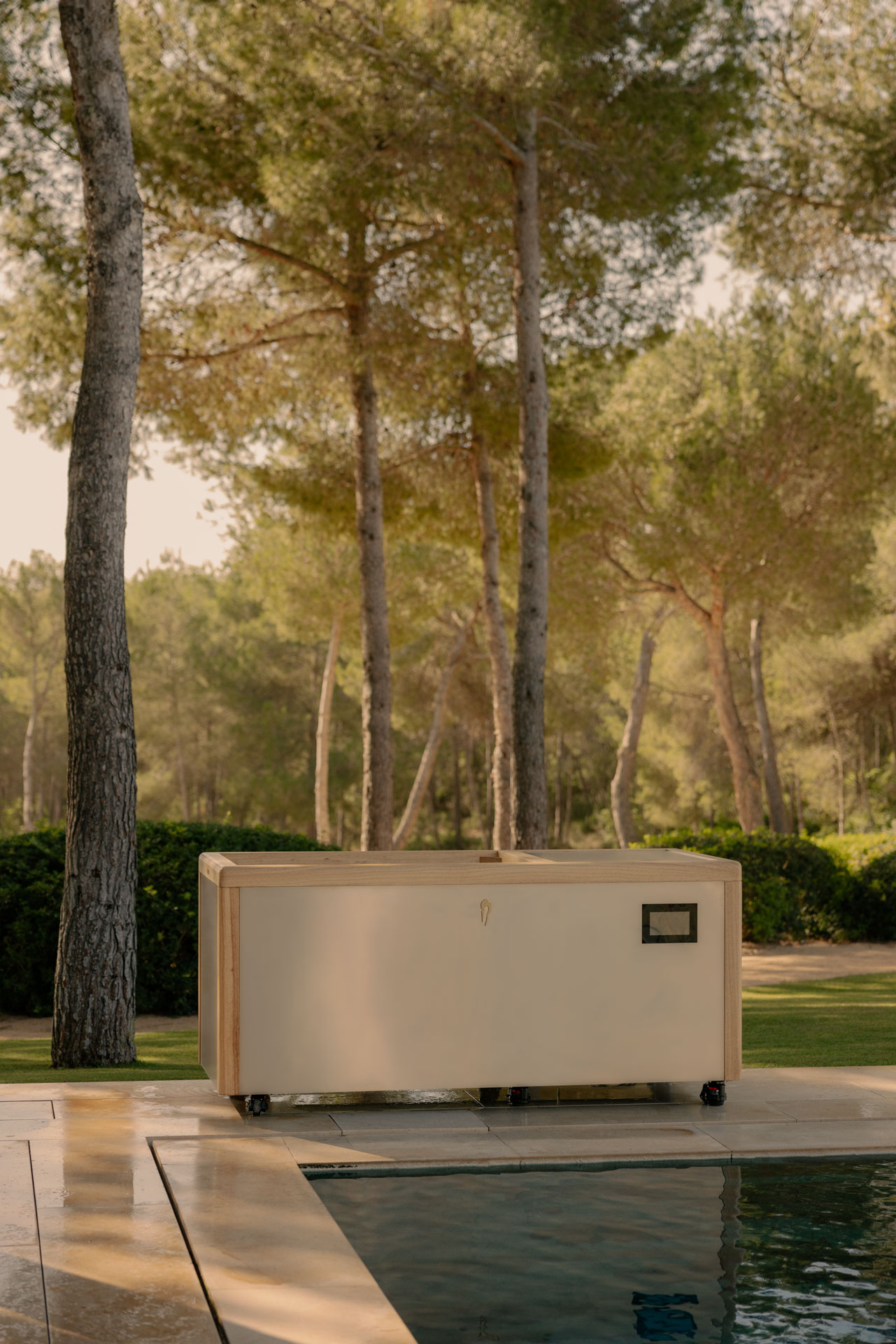 Wooden outdoor storage box on wheels beside a pool with pine trees in the background.
