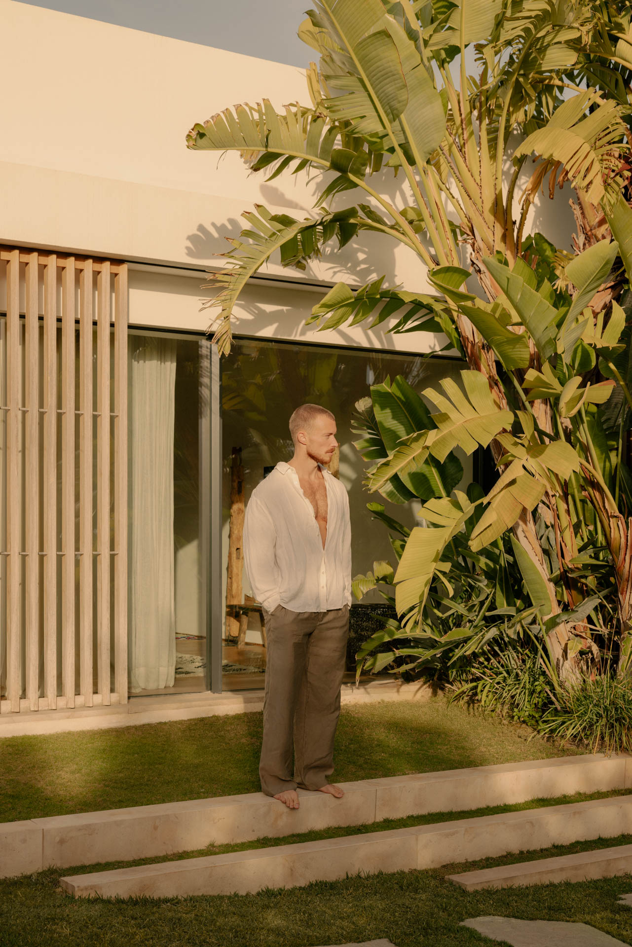 Barefoot man with short hair and beard wearing an open white shirt and brown pants standing on a stone ledge in front of a modern house and large tropical plants.