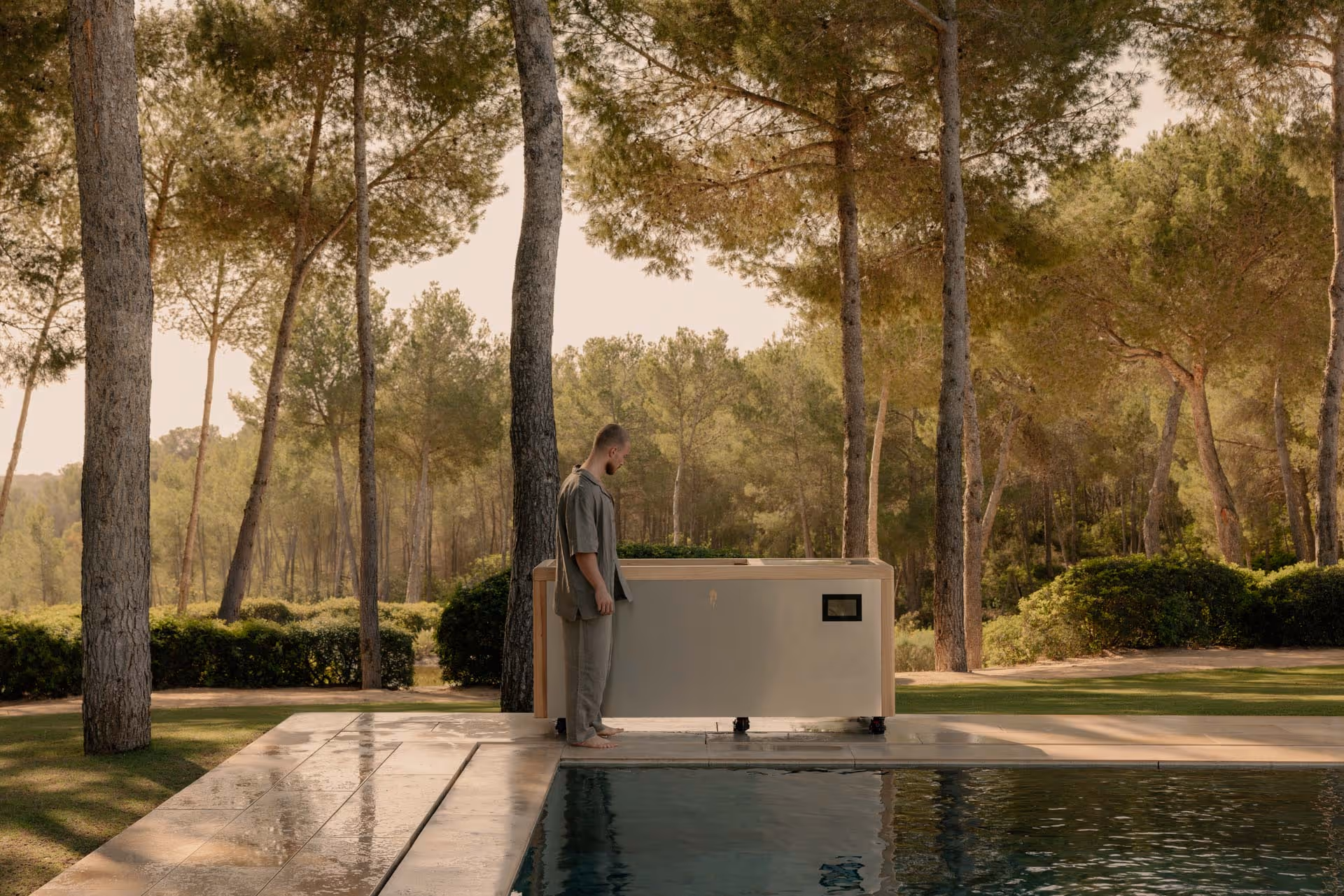 Man standing barefoot by a modern outdoor hot tub near a pool surrounded by trees.
