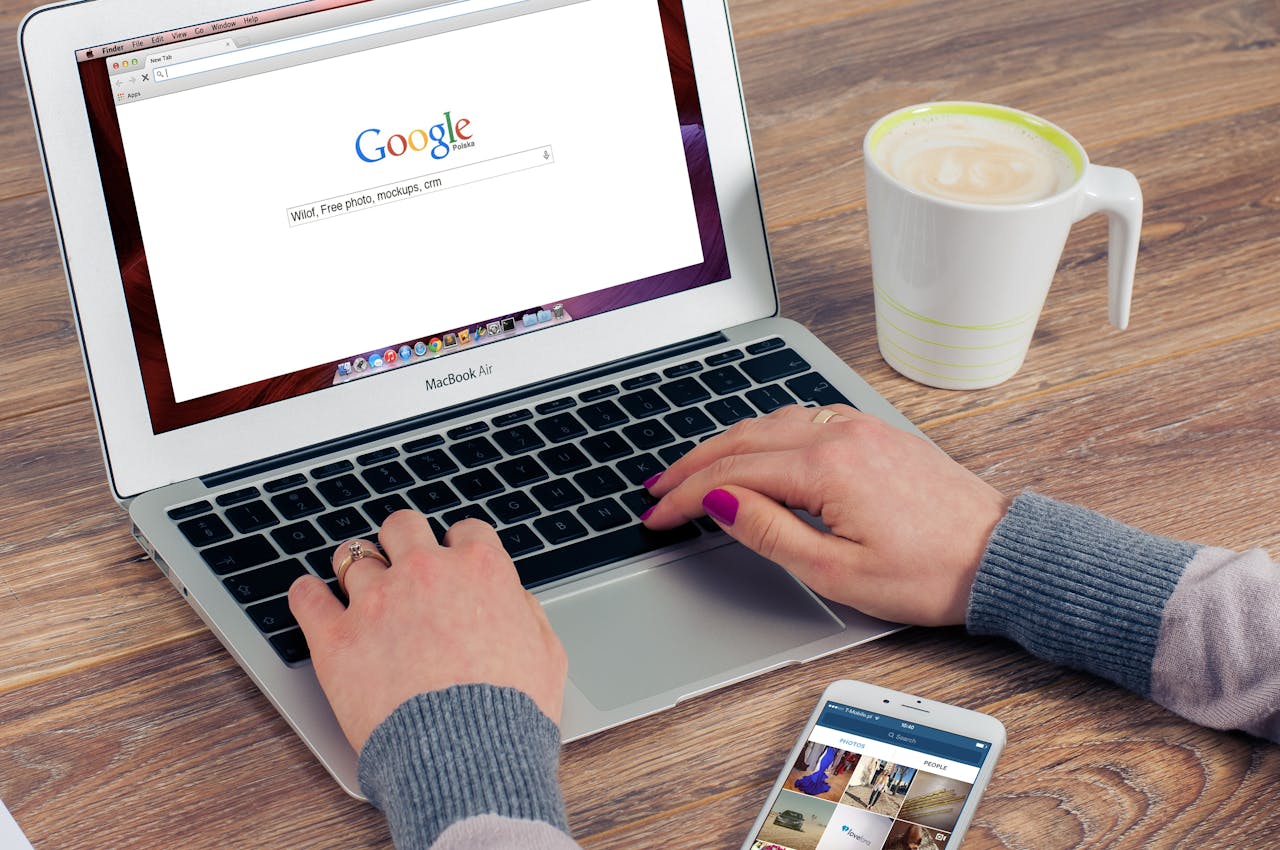 Person typing on a MacBook Air with a Google search page open, a cup of coffee, and a smartphone displaying a photo app on a wooden table.