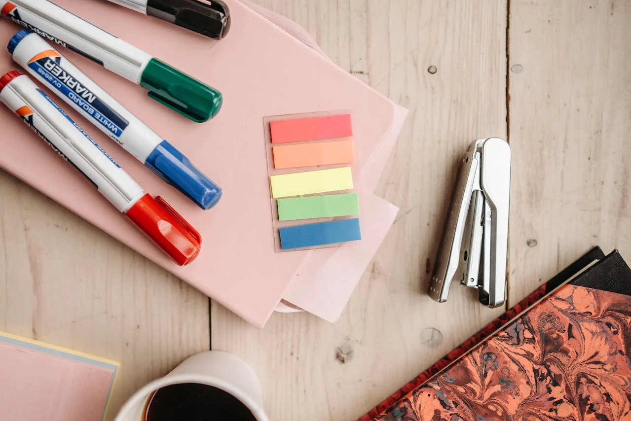 Top view of colorful whiteboard markers, pastel sticky tabs, a stapler, a marbled notebook, and a cup of black coffee on a wooden desk.