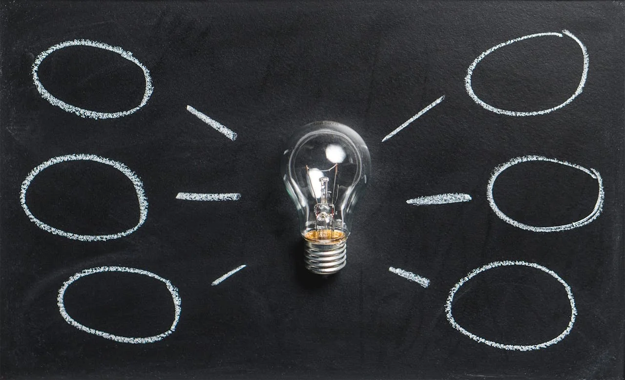 Light bulb in the center of a blackboard with six empty oval shapes connected around it by drawn lines, symbolizing brainstorming or idea mapping.
