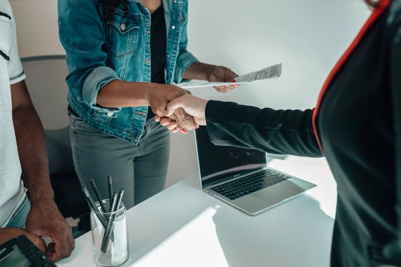 Two people shaking hands over a white desk with a laptop and a glass with pencils.