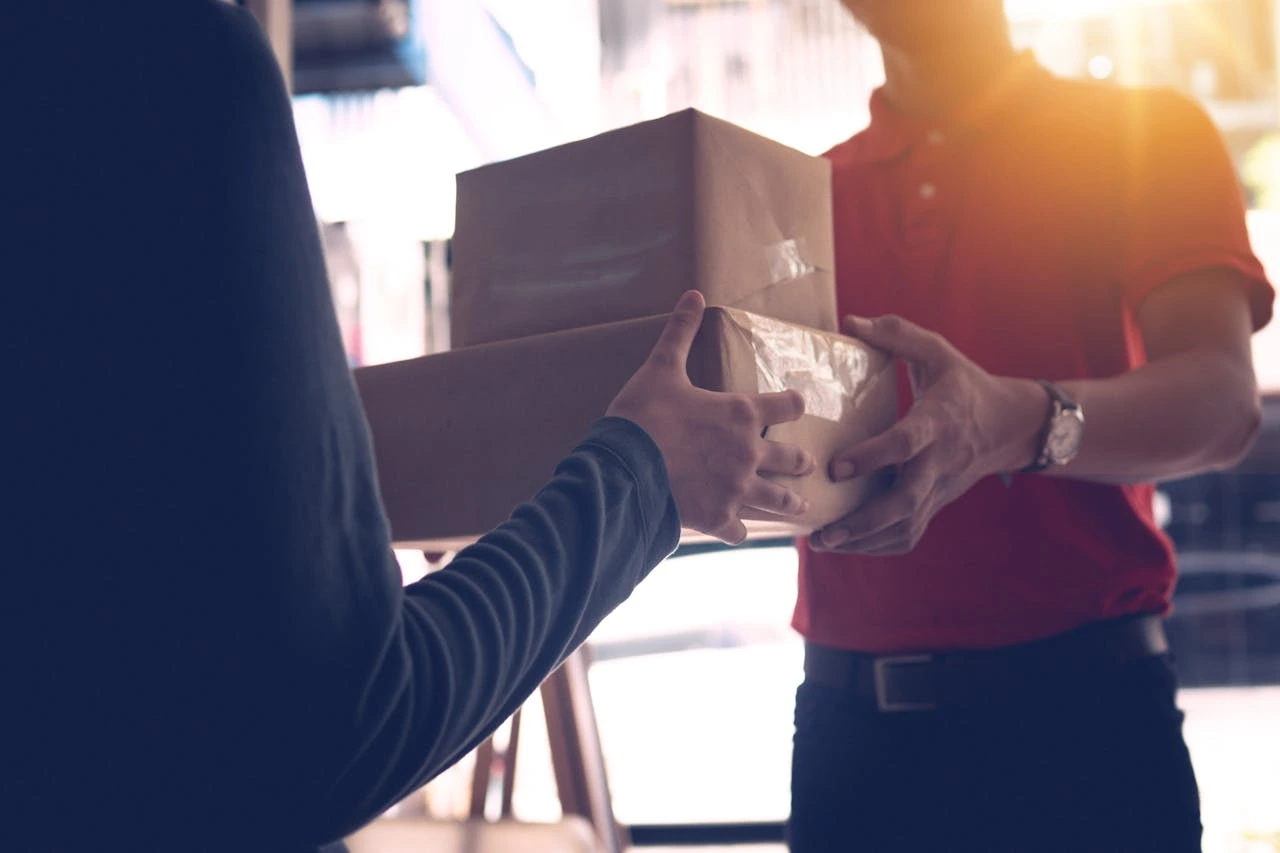 Person in red shirt handing two stacked cardboard boxes to another person wearing a dark long sleeve shirt.