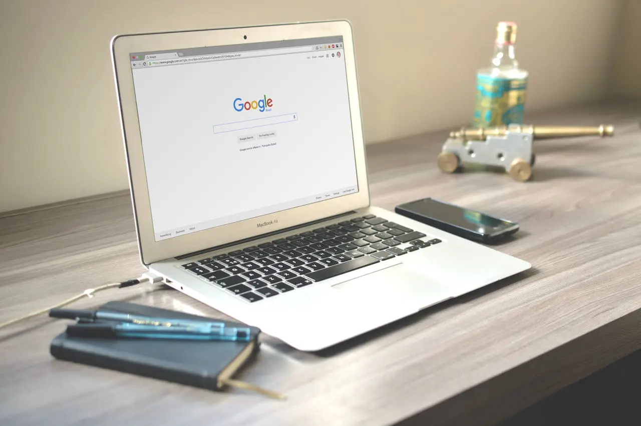 Open MacBook Air laptop on wooden desk displaying Google search page, with black smartphone, small notebook, two pens, and a decorative miniature cannon and bottle in the background.