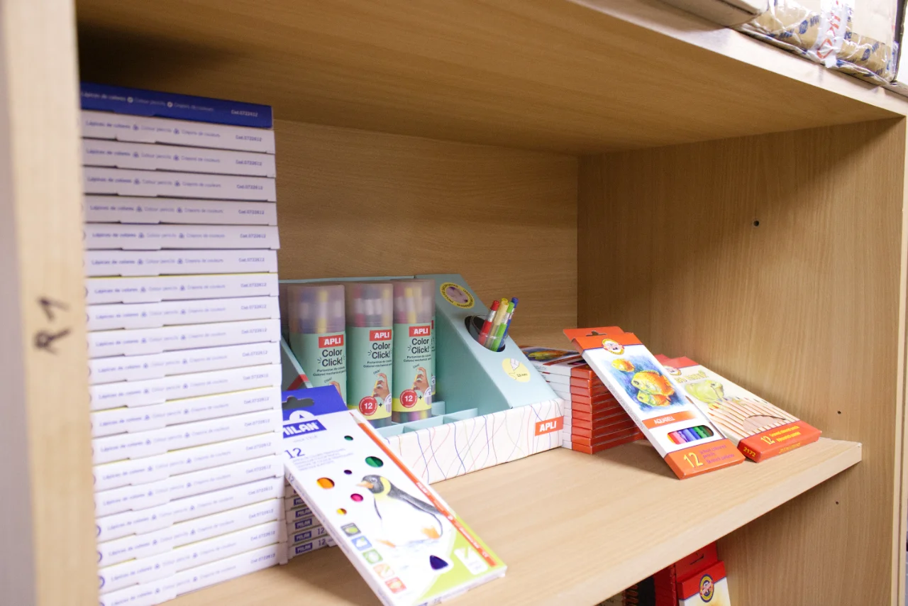 Wooden shelf stocked with stacks of white boxes, colorful marker packs, and assorted colored pencils and pens in packaging.