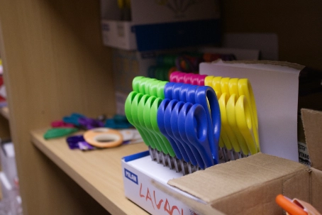 Rows of green, blue, and yellow scissors standing upright in a white box on a wooden shelf.
