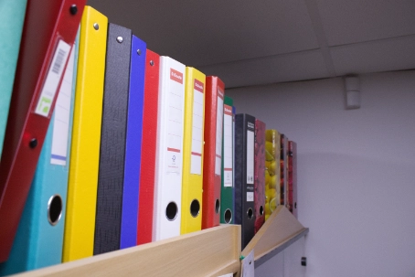 Close-up of colorful upright binders arranged on a wooden shelf against a plain wall.