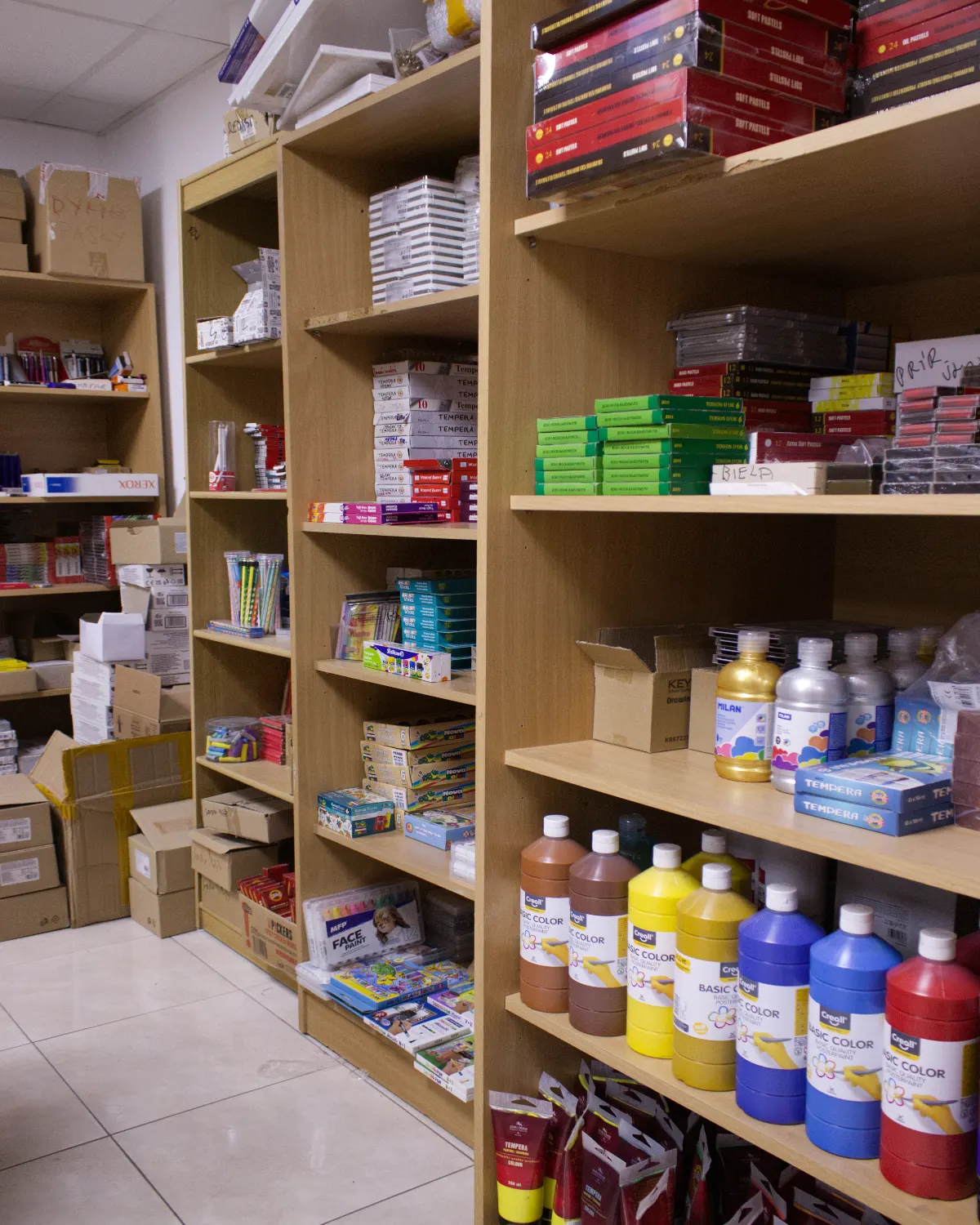 Shelves stocked with various art supplies including large bottles of paint, boxes of tempera colors, drawing materials, and boxed items in a storage room.