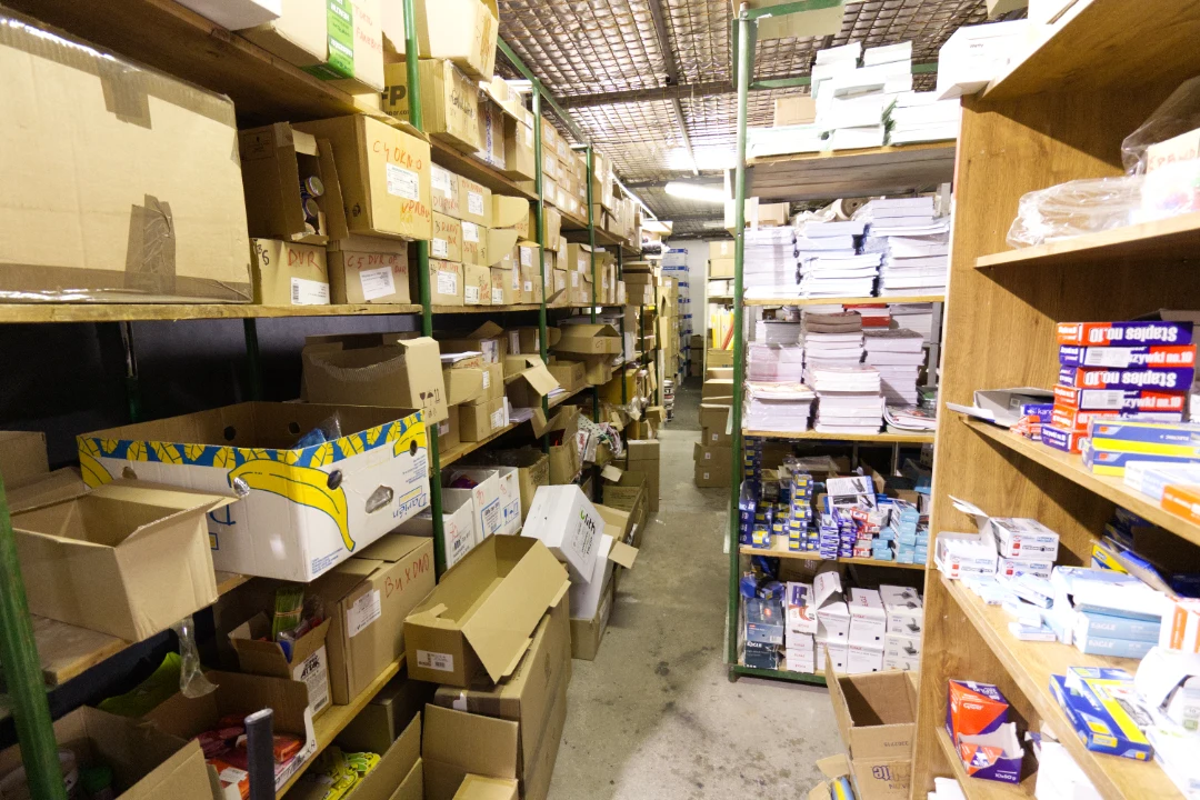 Narrow warehouse aisle lined with metal and wooden shelves filled with cardboard boxes and stacks of paper products.