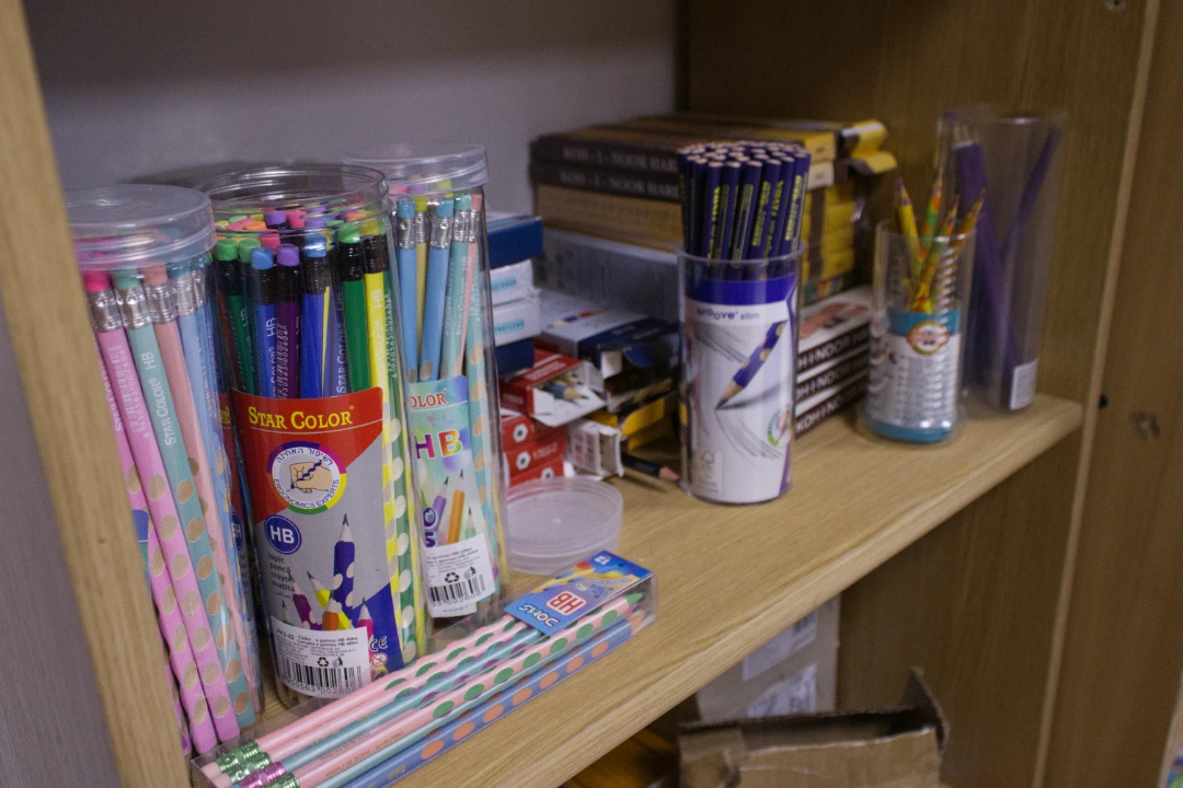 Wooden shelf with containers of colorful HB pencils, pens, erasers, and stacked boxes of school supplies.