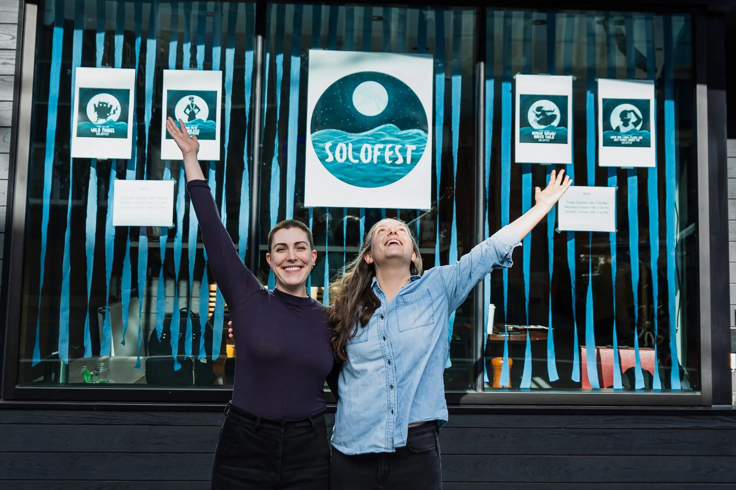 Two women celebrating in front of a Solofest sign at LunaSea Theatre Company, supported by CLG Injury Law in 2024 and 2025.
