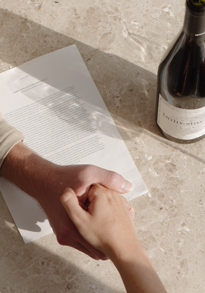 Two people holding hands over a contract on a marble table next to a wine bottle.