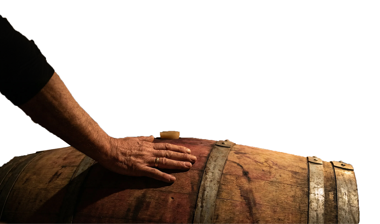 Hand with a wedding ring resting on a wooden barrel with metal bands against a black background.