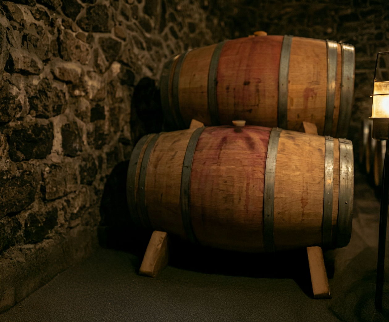 Two wooden barrels with metal hoops resting on wooden supports inside a dimly lit stone cellar.